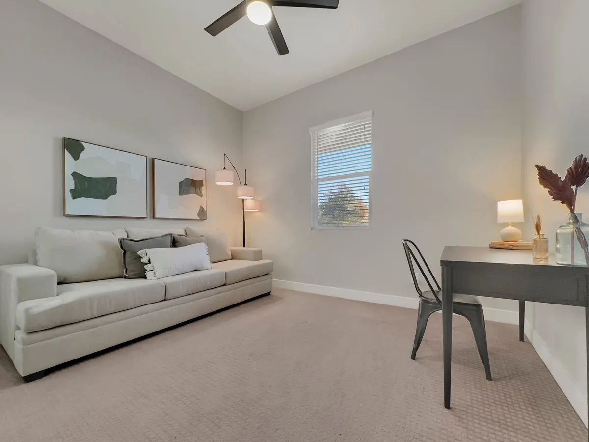 A dedicated office space featuring a sofa, desk with lamp, artwork on the wall, and a window with blinds. The space is serene, uncluttered, and designed for focus or relaxation.