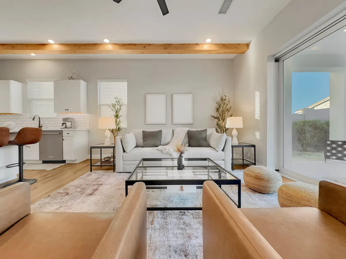 A cozy, open-concept living area with a plush sofa adorned with decorative pillows, a coffee table, and a spacious kitchen area visible in the background. Large glass doors let in abundant Arizona sunshine and lead to an outdoor space, reinforcing the sense of openness and connection.