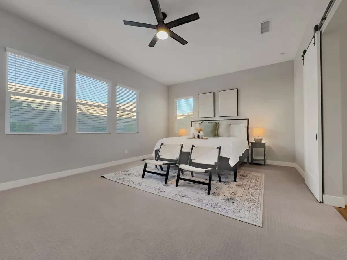 A spacious bedroom with a comfortable bed adorned with decorative pillows, two accent chairs at the foot of the bed, framed artwork on the wall, and a nightstand with a lamp. Sunlight filters in, enhancing the room’s airy, peaceful feel.