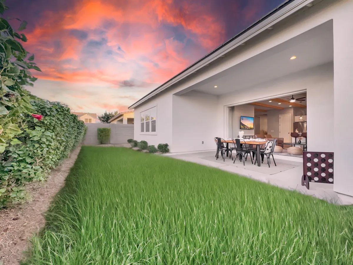 A lush green lawn stretches across a backyard, bordered by vibrant plantings. A spacious patio area is set with a dining table and chairs, inviting outdoor gatherings. The setting sun paints the sky in warm colors, casting a serene glow over the open space. The area feels welcoming, ideal for relaxation, playtime, or enjoying a meal outside.