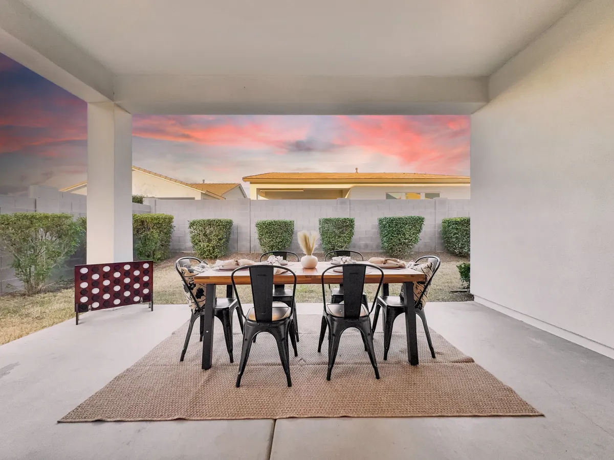 An inviting outdoor dining setup with a table and chairs on a decorative rug, all beneath a colorful sunset sky. The scene is warm and sociable, suggesting gatherings, al fresco meals, or peaceful evenings outdoors.