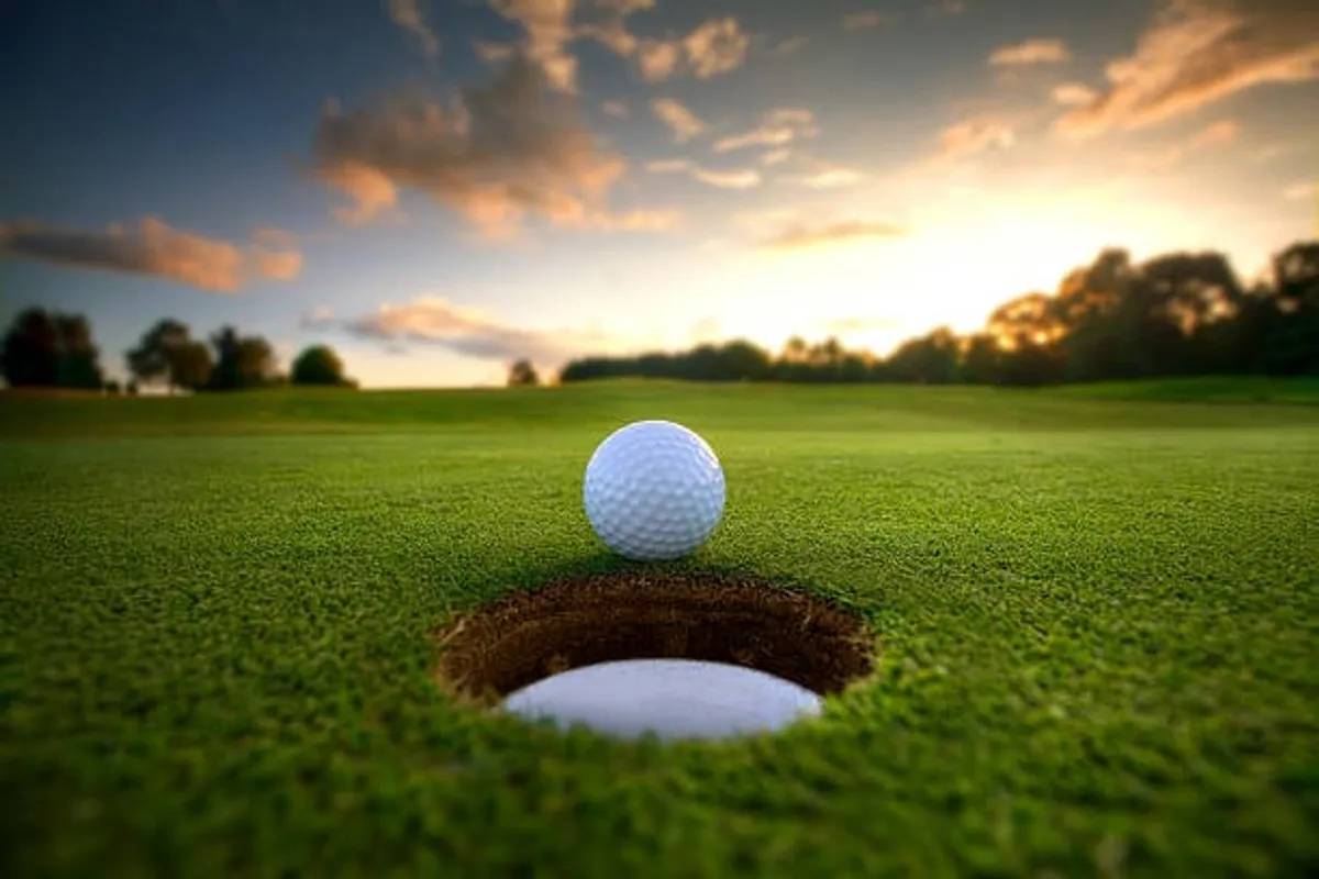 A close-up shot of a golf ball poised at the edge of a lush green hole, with the expansive golf course subtly visible in the background at sunset. The scene evokes both the excitement of play and the tranquil camaraderie found on the greens.