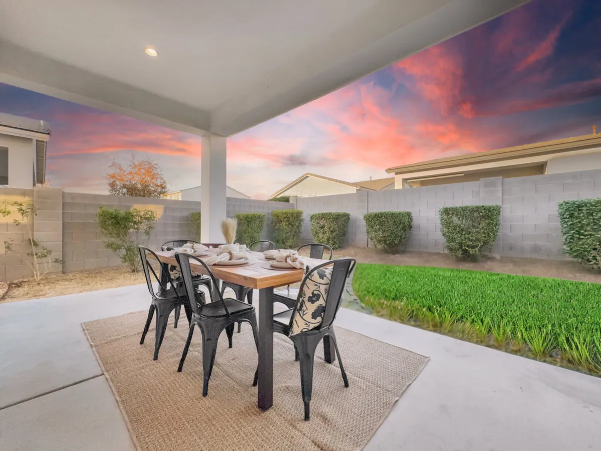 An inviting outdoor dining table set with tableware, surrounded by vibrant green grass under a colorful sunset sky. The scene is ready for a relaxed evening meal or social gathering in a tranquil backyard setting.